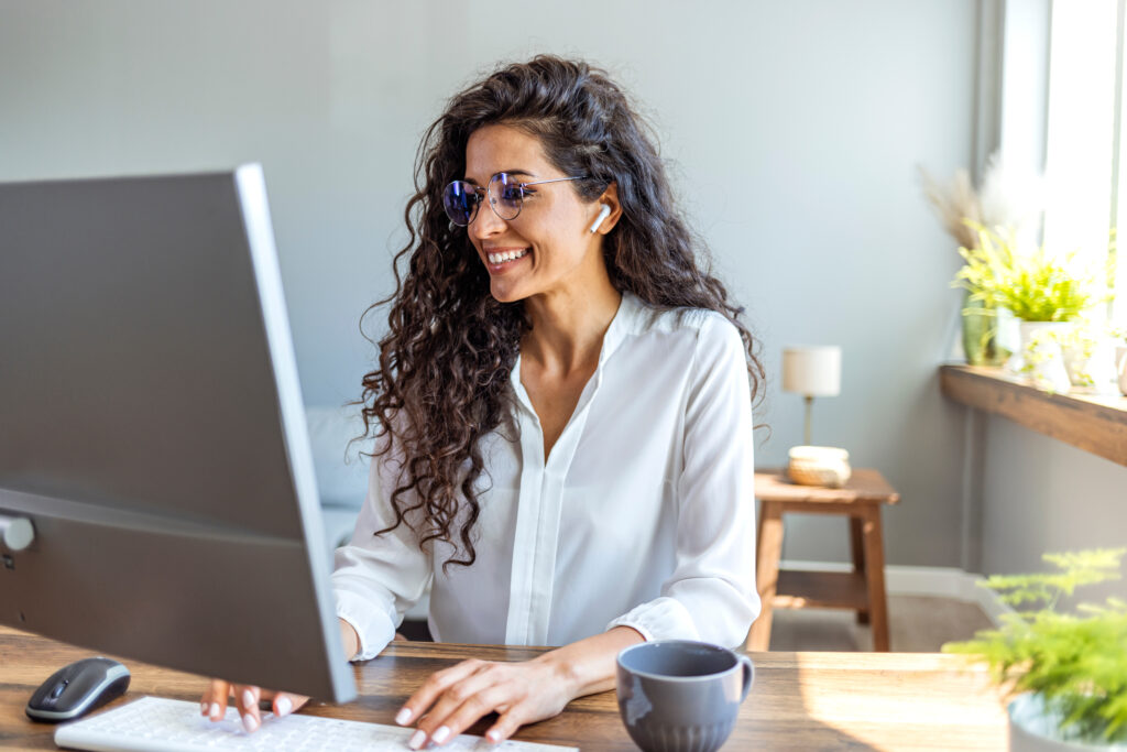Une femme souriante assise à son bureau dans un espace de coworking, effectuant une recherche sur son ordinateur portable.